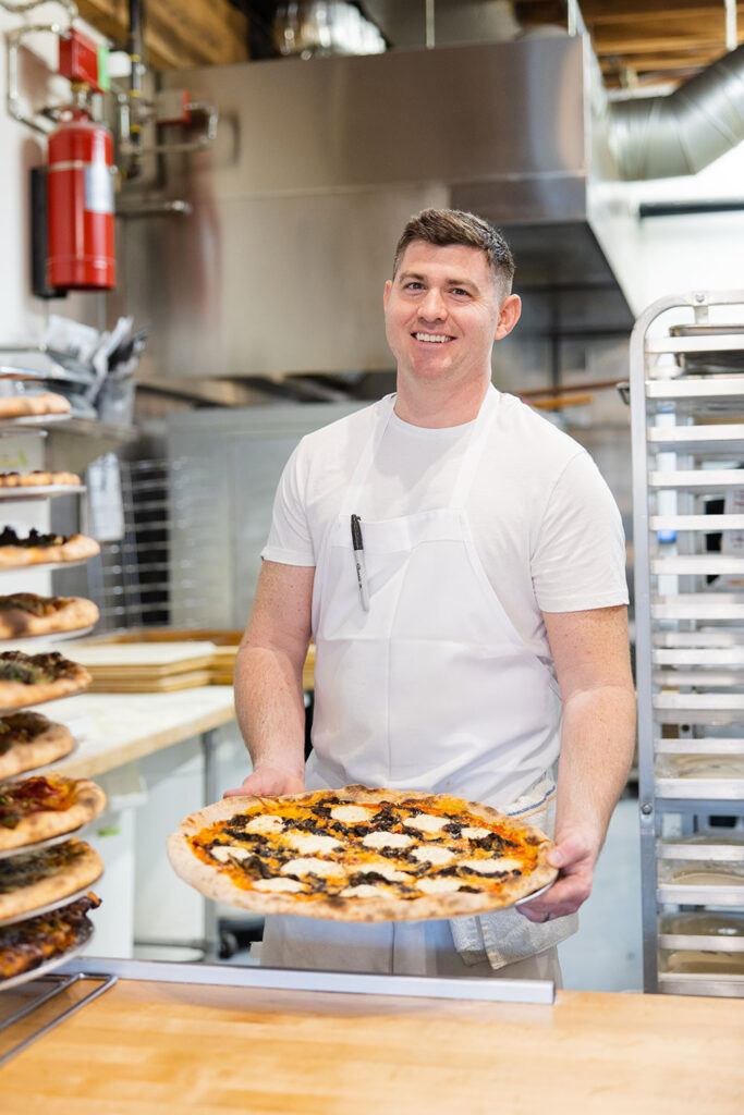 a pizza chef holds a freshly-made pizza and smiles at the pass at pizza thief