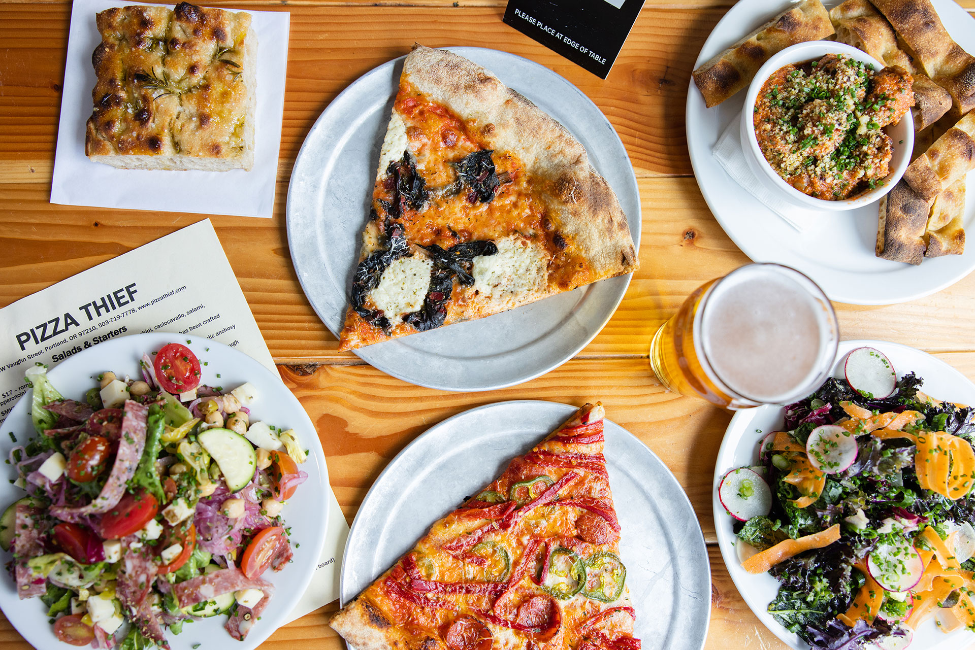 a sampling of pizza thief pizza slices, salads, beer, focaccia, and meatballs